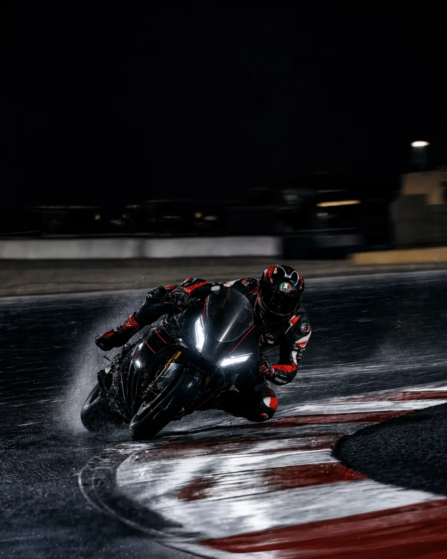 A motorcycle racer leans through a wet corner at night as water sprays from the tires.